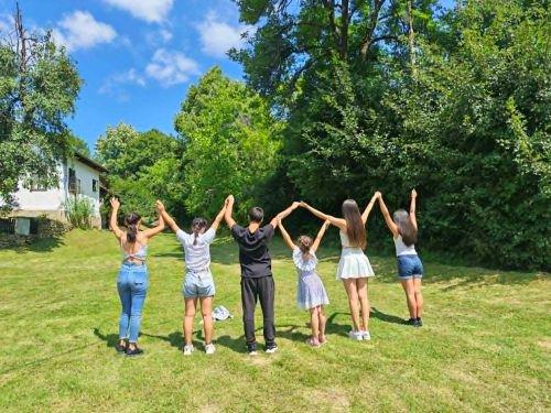 Seven children and young people standing in a line on the grass, arms raised and hands joined, seen from behind.