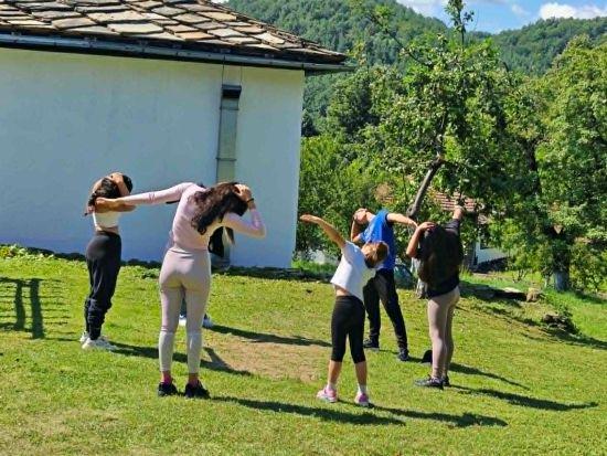 Children and young people stretching together on the grass beside a village house, wooded hills behind them.