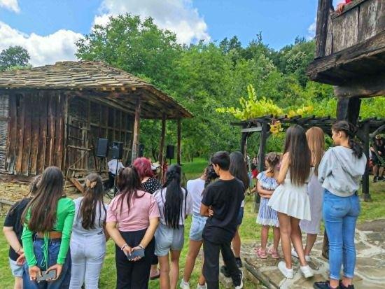 A group of children and young people watching a performance under a wooden shelter, seen from behind.